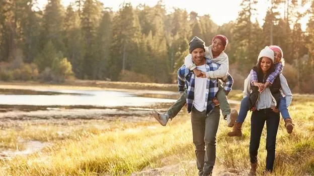 Parents carrying their children on their backs beside a lake and wooded landscape
